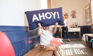 A playful kiddo holding a pillow in front of their face that says AHOY!