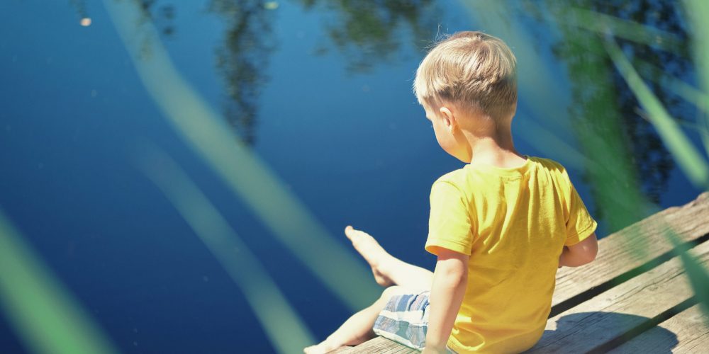 Happy child boy sitting on wooden pier near lake on summy summer day. Toddler kid in yellow t-shirt spend time on nature. Happy child boy sitting on wooden pier near lake on summy summer day. Toddler kid in yellow t-shirt spend time on nature.